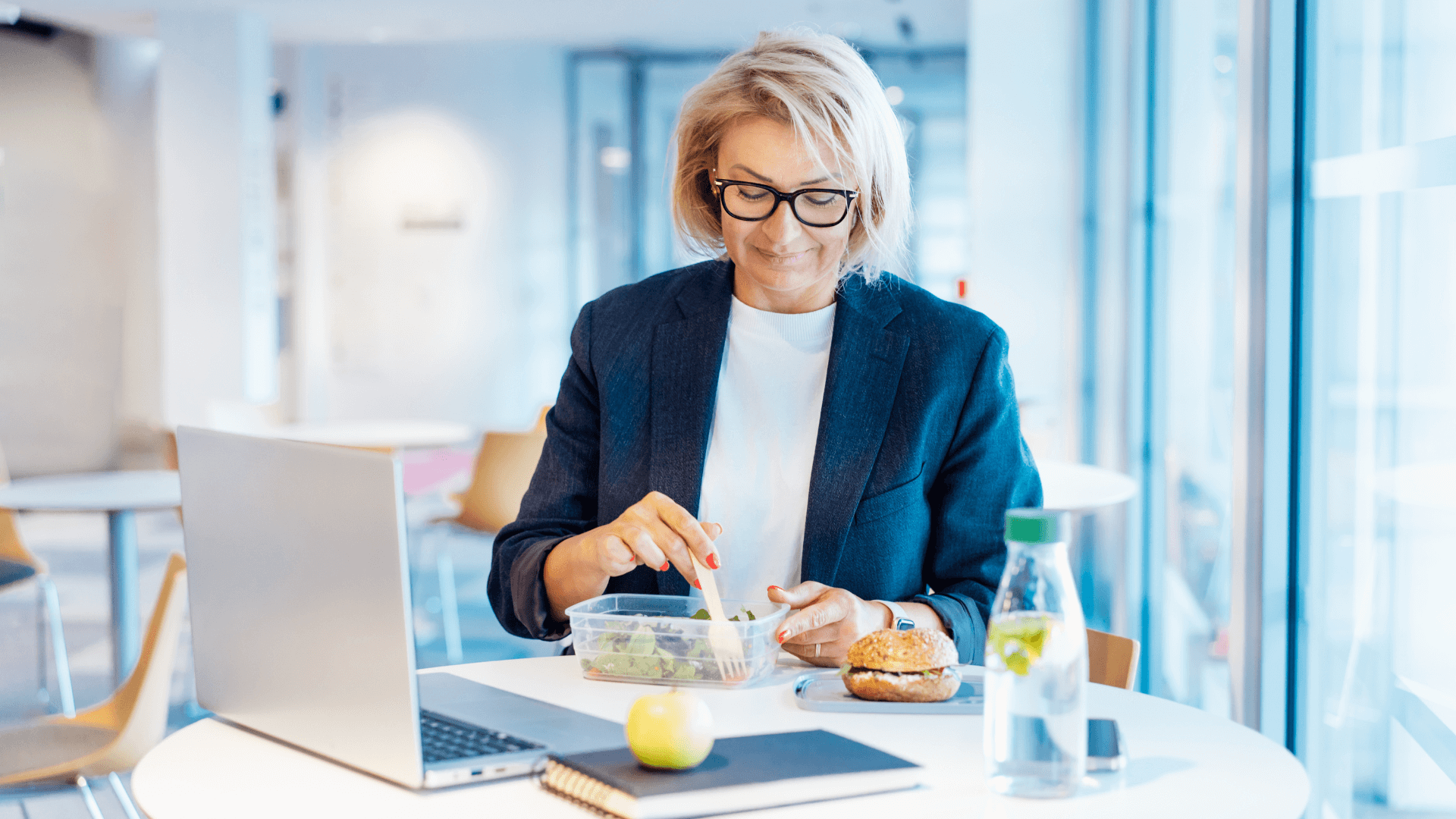 Eine Frau mit kurzen blonden Haaren und Brille, gekleidet in eine Businessjacke und einen weißen Pullover, isst einen Salat an ihrem Schreibtisch in einem hell erleuchteten Büro. Sie schaut auf einen Behälter mit Salat, den sie mit einer Gabel umrührt, während vor ihr ein Laptop, eine Flasche Wasser, ein Sandwich, ein Apfel und ein Notizbuch auf dem Tisch stehen.