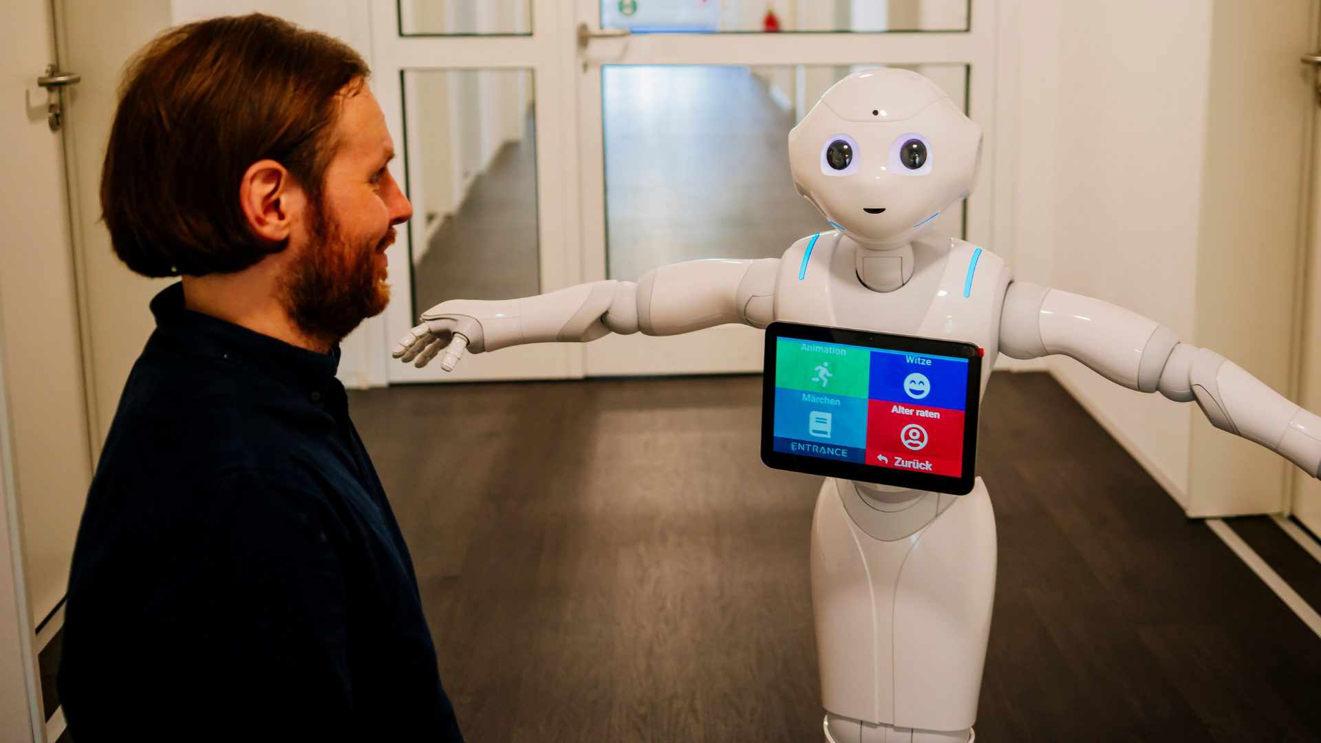 Robert Ranisch lächelt den humanoiden Roboter Pepper mit ausgestrecktem Arm in einem Flur an, auf dessen Bildschirm verschiedene interaktive Optionen angezeigt werden.